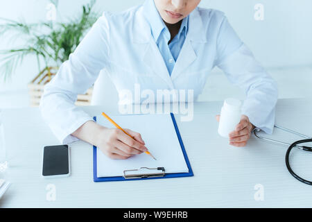 Cropped shot of young doctor writing on clipboard en étant assis sur le lieu de travail et la tenue de comprimés contenant Banque D'Images