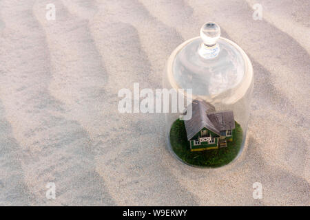 Maison en carton jouet avec pelouse verte protégée par un dôme en verre cloche parmi d'un désert de sable sans vie Banque D'Images