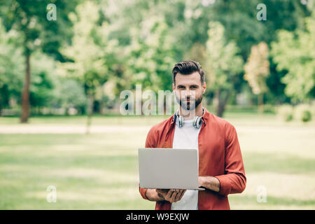 Young man standing in park with laptop in hands and looking at camera Banque D'Images
