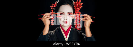 Belle geisha en kimono rouge avec des fleurs dans les cheveux maintenant près de baguettes yeux isolated on black, vue panoramique tourné Banque D'Images