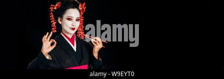 Smiling belle geisha en kimono noir avec des fleurs dans les cheveux rouge holding baguettes et montrant okay sign isolated on black, vue panoramique tourné Banque D'Images