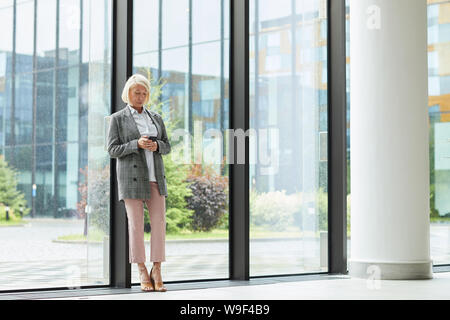 Serious mature woman in stylish costume formel s'appuyant sur la grande fenêtre et en utilisant son téléphone portable au bureau moderne Banque D'Images