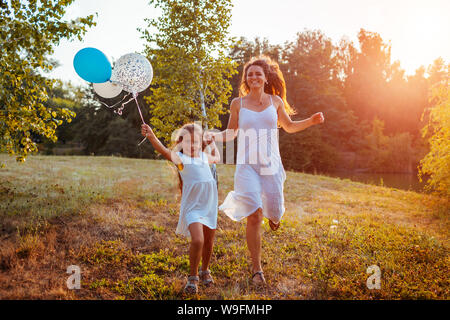 Happy little girl tournant avec la mère et la tenue en main. baloons Family in parc d'été au coucher du soleil. Journée internationale de Childrens Banque D'Images