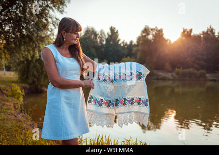 Mature Woman holding serviette avec broderie traditionnelle ukrainienne en face de river in autumn Park au coucher du soleil. Senior woman est fier de sa nationalité Banque D'Images