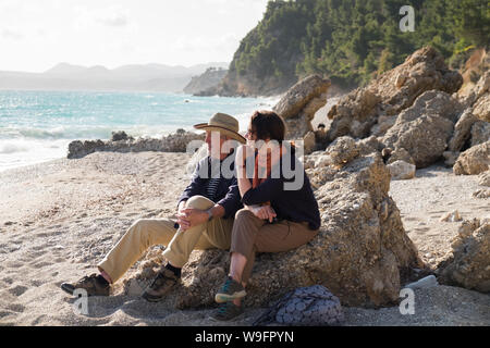 Un couple dans la fin des années 60 début des années 70, s'asseoir et admirer la vue sur la plage isolée et vide Paradissi à Kefalonia, Grèce. Banque D'Images