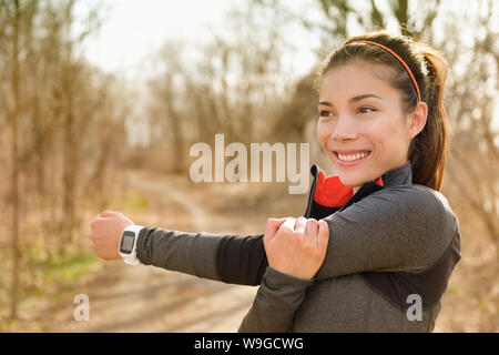 Femme fitness stretching arms avec smartwatch avant d'exécuter ou d'entraînement cardio. Happy Asian girl doing échauffement avant la course avec moniteur de fréquence cardiaque à l'extérieur parc au cours de l'automne. Banque D'Images