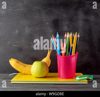 Vert pomme, banane mûre sur une pile de carnets, papeterie rouge verre avec multi-couleur crayons en bois sur l'arrière-plan d'un boa craie noire vide Banque D'Images