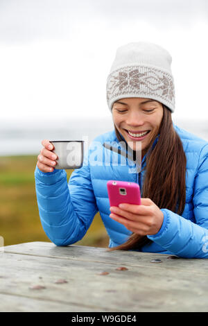 Femme à l'extérieur à l'aide de smart phone app de boire du café à partir de la Tasse thermos assis dehors chaud porter Down jacket et Knit hat. Pretty young mixed race woman chinois asiatique dans la vie active. Banque D'Images