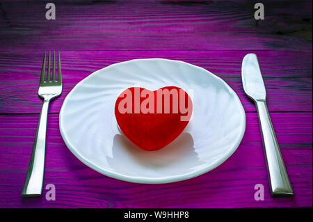 Peluche coeur rouge sur une plaque de métal blanc, fourchette et couteau sur table en bois violet foncé. Banque D'Images