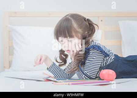 Cute little girl doing homework, écriture et dessin avec des crayons de couleur sur le lit chez lui. Banque D'Images