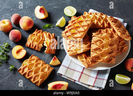 Mini tartes pâte feuilletée avec de la confiture de pêches et de lime garniture sur une assiette blanche sur une table en béton avec des pêches fraîches, les feuilles de menthe et de tranches de lime, horizontal Banque D'Images