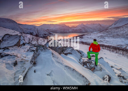 Randonneur admire la mer gelée entouré par la neige encadré par le ciel orange au coucher du soleil, de Torsken, Senja, Troms County, arctique, Norway, Scandinavia, Europe Banque D'Images