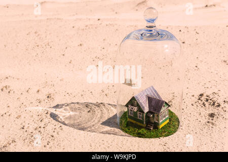 Maison en carton jouet avec pelouse verte protégée par un dôme en verre cloche parmi d'un désert de sable sans vie Banque D'Images