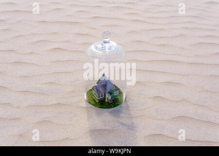 Maison en carton jouet avec pelouse verte protégée par un dôme en verre cloche parmi d'un désert de sable sans vie Banque D'Images