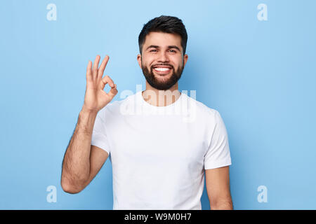 Bel homme barbu positive en T-shirt blanc isolé sur fond bleu indiquant ok sign.close up portrait, langue de corps, tout est ok, allrigh Banque D'Images