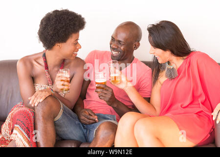 Trois amis multiculturels, rire et parler sur la table avec les boissons alcoolisées lors d'une partie de la chambre ou rassemblement Banque D'Images