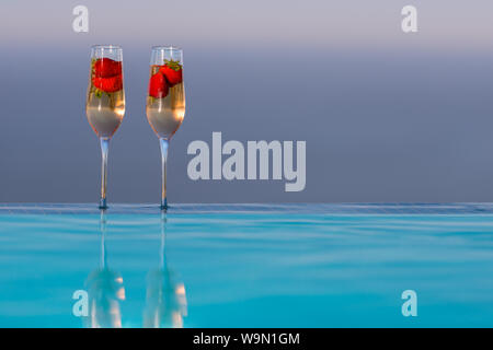 Deux verres de champagne avec des fraises sur le bord de piscine à débordement coucher du soleil derrière eux Banque D'Images