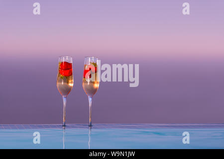 Deux verres de champagne avec des fraises sur le bord de piscine à débordement coucher du soleil derrière eux Banque D'Images