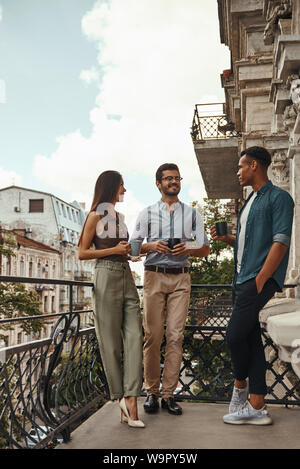 Avoir une pause-café. Les jeunes collègues gais dans des vêtements décontractés et de parler avec les tasses holding les uns les autres tout en se tenant sur le balcon. Les collègues à se reposer. La vie de bureau Banque D'Images