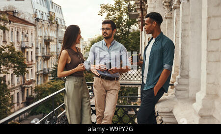 La conversation. Les jeunes collègues gais dans des vêtements décontractés et de parler avec les tasses holding les uns les autres tout en se tenant sur le balcon. Les collègues à se reposer. Pause café Banque D'Images