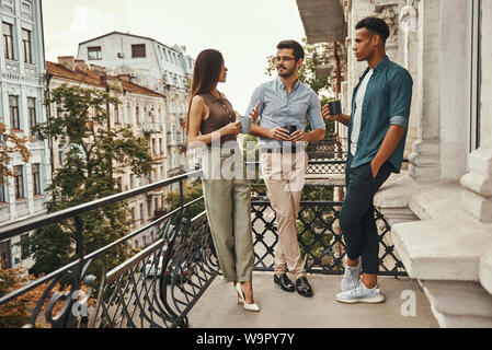 La vie de bureau. Les jeunes collègues gais dans des vêtements décontractés et de parler avec les tasses holding les uns les autres tout en se tenant sur le balcon. Les collègues à se reposer. Pause café Banque D'Images