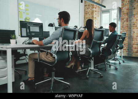 Tâches courantes. Vue arrière de jeunes employés travaillant sur les ordinateurs alors que sitting at desk in modern espace ouvert. Concept d'emploi. Lieu de travail Banque D'Images