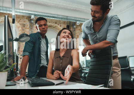 L'examen du projet. Groupe de jeunes et joyeux trois personnes dans les tenues de l'analyse des données et souriant tout en travaillant dans un bureau moderne. Concept d'entreprise. Lieu de travail Banque D'Images