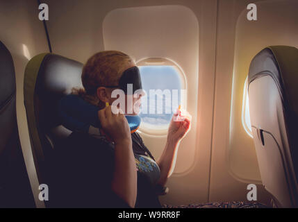 Femme de 33 ans voyageant avec un oreiller, un masque de sommeil et boules quiès en cabine avion sur siège passager, voler avec l'avion, un voyage confortable confortable Banque D'Images