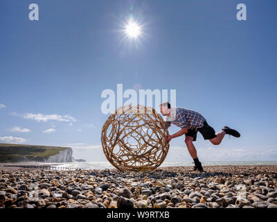 Cuckmere Haven, au Royaume-Uni. 15 août, 2019. Artiste Keith Pettit place le premier d'une série de machine à vapeur-bent chestnut sphères sur la plage de Cuckmere Haven dans l'East Sussex. Faites en collaboration avec trug bouilloire Charlie Groves, les formes complexes sont inspirés par les ossements de coccolithes ancienne - le carbonate de calcium reste qui composent la craie des South Downs. L'exposition 'What Lies Beneath' sera à l'emblématique coastguard cottages donnant sur les falaises des sept Sœurs (photo). Crédit : Jim Holden/Alamy Live News Banque D'Images