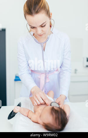 Très belle jeune femme blonde médecin pédiatre examine baby girl with stethoscope contrôle du rythme cardiaque. bébé est posé sur la balance. Banque D'Images