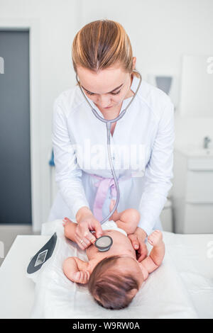 Très belle jeune femme blonde médecin pédiatre examine baby girl with stethoscope contrôle du rythme cardiaque. bébé est posé sur la balance. Banque D'Images