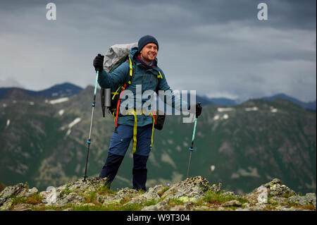 Randonneur souriant homme se sur la falaise contre green mountain range. L'homme a un sac à dos et bâtons de marche. Les randonnées en montagne. Copy space Banque D'Images