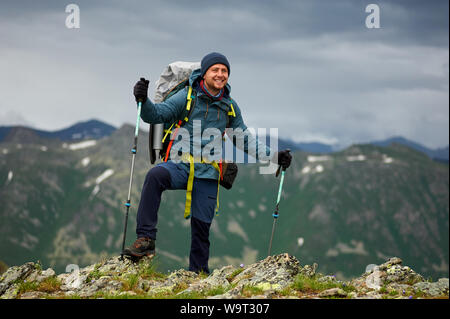 Randonneur souriant homme se sur la falaise contre green mountain range. L'homme a un sac à dos et bâtons de marche. Les randonnées en montagne. Copy space Banque D'Images