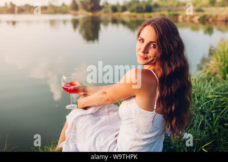 Jeune femme bénéficiant d'un verre de vin sur les bords de la rivière au coucher du soleil. Woman admiring paysage tout en ayant un verre dans le parc d'été Banque D'Images