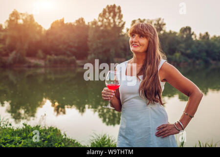 Mature Woman enjoying verre de vin par river in autumn park. Senior woman paysage admire ayant verre Banque D'Images