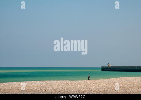 Une personne qui marche sur la plage de galets à Dieppe, en France, sur un jour de printemps ensoleillé mais venteux. Le traversier brise-lames pour la droite. Banque D'Images