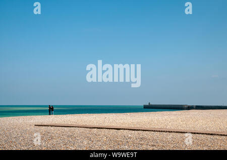 Deux personnes en pause sur la plage de galets à Dieppe, en France, sur un jour de printemps ensoleillé mais venteux. Le traversier brise-lames pour la droite. Banque D'Images