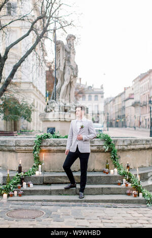 Groom smiling élégant dans un manteau gris, posant à l'extérieur, debout sur les marches en pierre du monument dans le vieux centre-ville avant la cérémonie de mariage. Banque D'Images