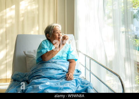 Asian woman sitting and holding glass of water au lit à l'hôpital. Banque D'Images
