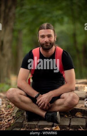 Portrait de jeune homme barbu dans la lumière naturelle tout en vous reposant dans le parc dans les escaliers. Homme de race blanche à l'athlète à l'appareil photo Banque D'Images