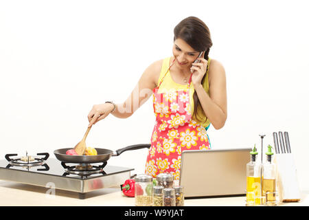 Jeune femme cuisant des aliments dans une poêle tout en parlant téléphone portable Banque D'Images