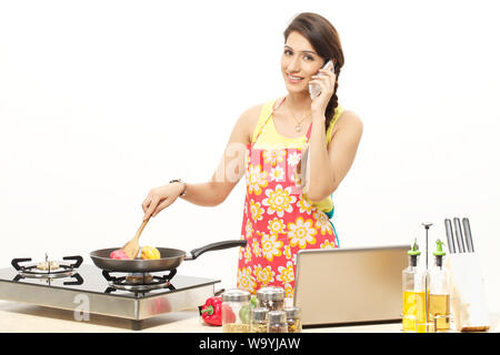 Jeune femme cuisant des aliments dans une poêle tout en parlant téléphone portable Banque D'Images