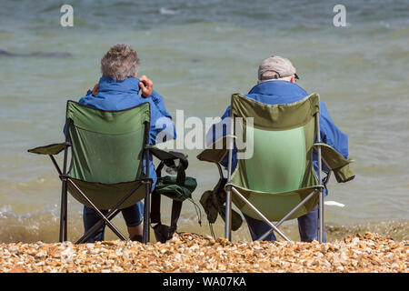 un couple ou un mari et une femme âgés à la retraite assis dans des chaises pliables sur la plage au bord de la mer portant des manteaux et des chapeaux. Banque D'Images