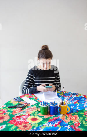 Jeune femme artiste de talent de taper un message sur portable assis à table avec une serviette fleuri dans son studio avec fond blanc. Utilise des crayons, papier, Banque D'Images