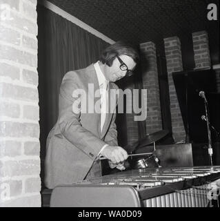 Années 1960, historique, un musicien en costume et cravate avec laine de bois recouvert de maillets dans ses mains jouant le marimba dans un lieu intérieur, Londres, Angleterre, Royaume-Uni. Le marimba, un instrument de percussion semblable à le xylophone, dispose d'un ensemble de barres de bois que lorsque frappé produisent des sons musicaux. Les tuyaux suspendus sous les barres ampify le son. Banque D'Images