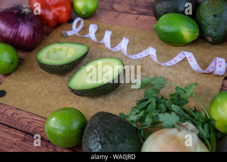 Moitié avocado guacamole ingrédients sur planche à découper en bois avec ruban à mesurer. La coriandre fraîche, l'oignon, limes et poivron rouge. Régime alimentaire sain matières f Banque D'Images