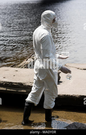 Vue de côté de l'inspecteur de l'eau en costume de protection et un masque respiratoire holding tubes à essai et la fiole à l'eau des échantillons au river Banque D'Images
