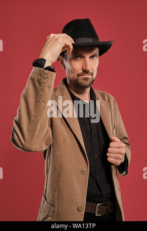 Studio shot of un homme avec barbe et moustache, porte manteau brun, en touchant son chapeau et regardant la caméra tout en se posant contre Banque D'Images