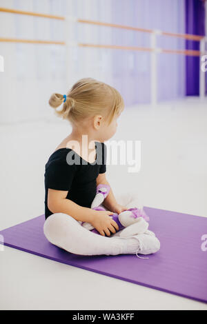 Jolie petite blonde ballerine fille assise sur un tapis mauve dans une classe de ballet et de faire de l'exercice, fond blanc. Banque D'Images