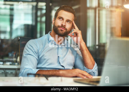 La réflexion sur l'entreprise. Jeune homme barbu dans l'usure formelle de toucher sa tête, souriant et à l'écart tout en travaillant dans un bureau moderne. Concept d'emploi. Lieu de travail Banque D'Images